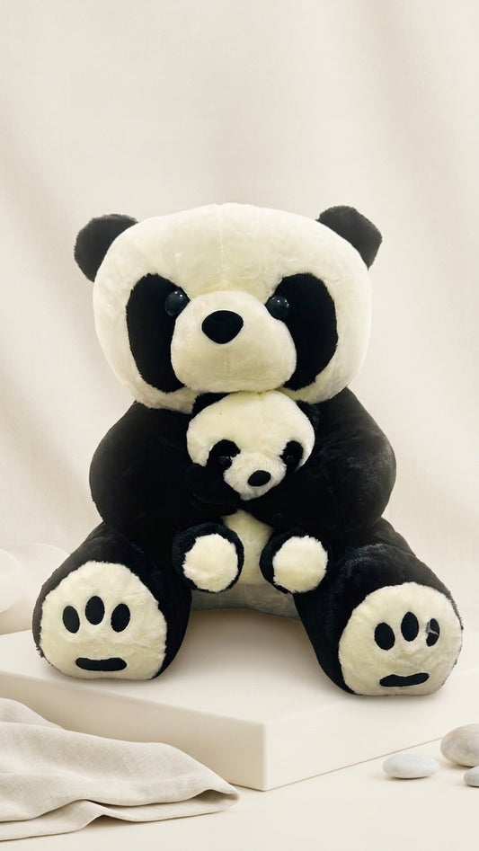 A black and white plush panda toy sitting on a white pedestal and holding a smaller matching panda, photographed in a clean studio setting with soft neutral fabric and small stones.
